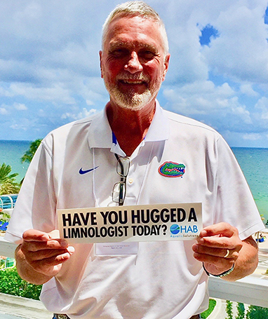 photo of a lakewatch director Mark Hoyer holding a bumper sticker that reads have you hugged a limnologist today